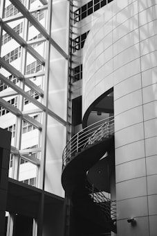A modern architectural space with geometric shapes, featuring a spiral staircase encased in glass and metal. The building's exterior is adorned with large, reflective windows that accentuate the sleek, contemporary design.