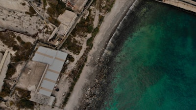 Aerial view of a coastal area featuring an industrial complex next to a beach. The complex includes several large buildings and infrastructure with pathways and possible machinery. The shoreline consists of rocky terrain leading into clear turquoise water.