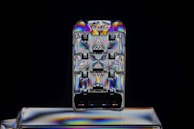 A small rectangular, transparent object featuring a swirling pattern of vibrant, iridescent colors. It appears to be a close-up of a plastic USB connector or similar electronic component against a dark background, creating a stark contrast. The colors shift across the surface, showing blues, purples, and yellows in a symmetrical layout.