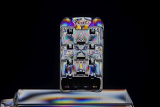 A small rectangular, transparent object featuring a swirling pattern of vibrant, iridescent colors. It appears to be a close-up of a plastic USB connector or similar electronic component against a dark background, creating a stark contrast. The colors shift across the surface, showing blues, purples, and yellows in a symmetrical layout.