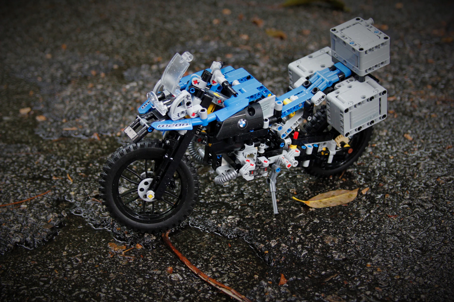 A blue and white mechanical motorcycle model with gray panniers standing on wet asphalt