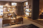 A luxurious shoe store interior with wooden walls and shelves displaying various designer shoes. There are comfortable leather chairs and a small wooden table placed in the center. The room has warm lighting and a clean, organized arrangement, creating a sophisticated atmosphere.