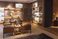A luxurious shoe store interior with wooden walls and shelves displaying various designer shoes. There are comfortable leather chairs and a small wooden table placed in the center. The room has warm lighting and a clean, organized arrangement, creating a sophisticated atmosphere.