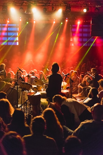 A live music performance with a drummer at the center stage, surrounded by a large audience. The scene is illuminated by vibrant red and orange stage lights, creating a lively atmosphere. Several musicians with brass instruments are also visible on the right side.