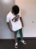 A person is standing indoors wearing a white t-shirt featuring a colorful graphic design with the word 'PYRAMID' and a cartoon character. They are also wearing green pants with red stripes and black sneakers with a checkered pattern. The person is looking downwards.