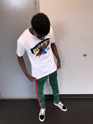 A person is standing indoors wearing a white t-shirt featuring a colorful graphic design with the word 'PYRAMID' and a cartoon character. They are also wearing green pants with red stripes and black sneakers with a checkered pattern. The person is looking downwards.