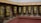 A series of traditional prayer wheels are spinning, mounted on a wooden frame against a colorful, intricately painted wall depicting various religious figures and motifs. The wheels have golden inscriptions and designs on a dark background, creating a rhythmic blur as they spin.