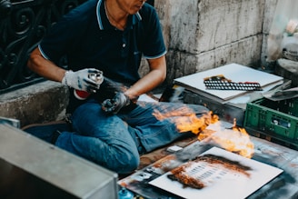 A person sits cross-legged on a pavement, using a spray paint can while creating street art. Flames are present on a piece of paper in front of them, indicating a dramatic or bold technique being used. Various art supplies and additional paper sheets are scattered around, revealing the setting is likely outdoors and informal.