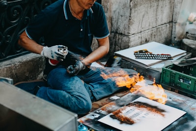 A person sits cross-legged on a pavement, using a spray paint can while creating street art. Flames are present on a piece of paper in front of them, indicating a dramatic or bold technique being used. Various art supplies and additional paper sheets are scattered around, revealing the setting is likely outdoors and informal.