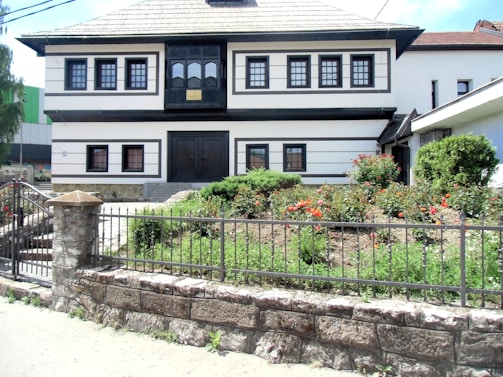A two-story white building with black trim details featuring multiple small windows and a large wooden door at the front. In front of the building, a well-maintained garden is visible, with a variety of red and orange roses and green shrubs. A stone fence with metal railings encloses the garden area.
