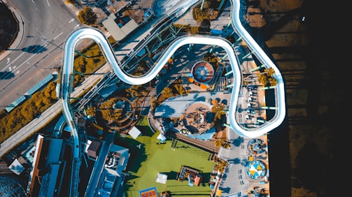 An aerial view of an amusement park featuring various rides and structures. A prominent white roller coaster track winds its way through the scene. Surrounding the roller coaster are other attractions such as a circular ride with a colorful roof, various pathways, and smaller attractions. Adjacent to the amusement park, a road with cars and sidewalks can be seen, along with some buildings.