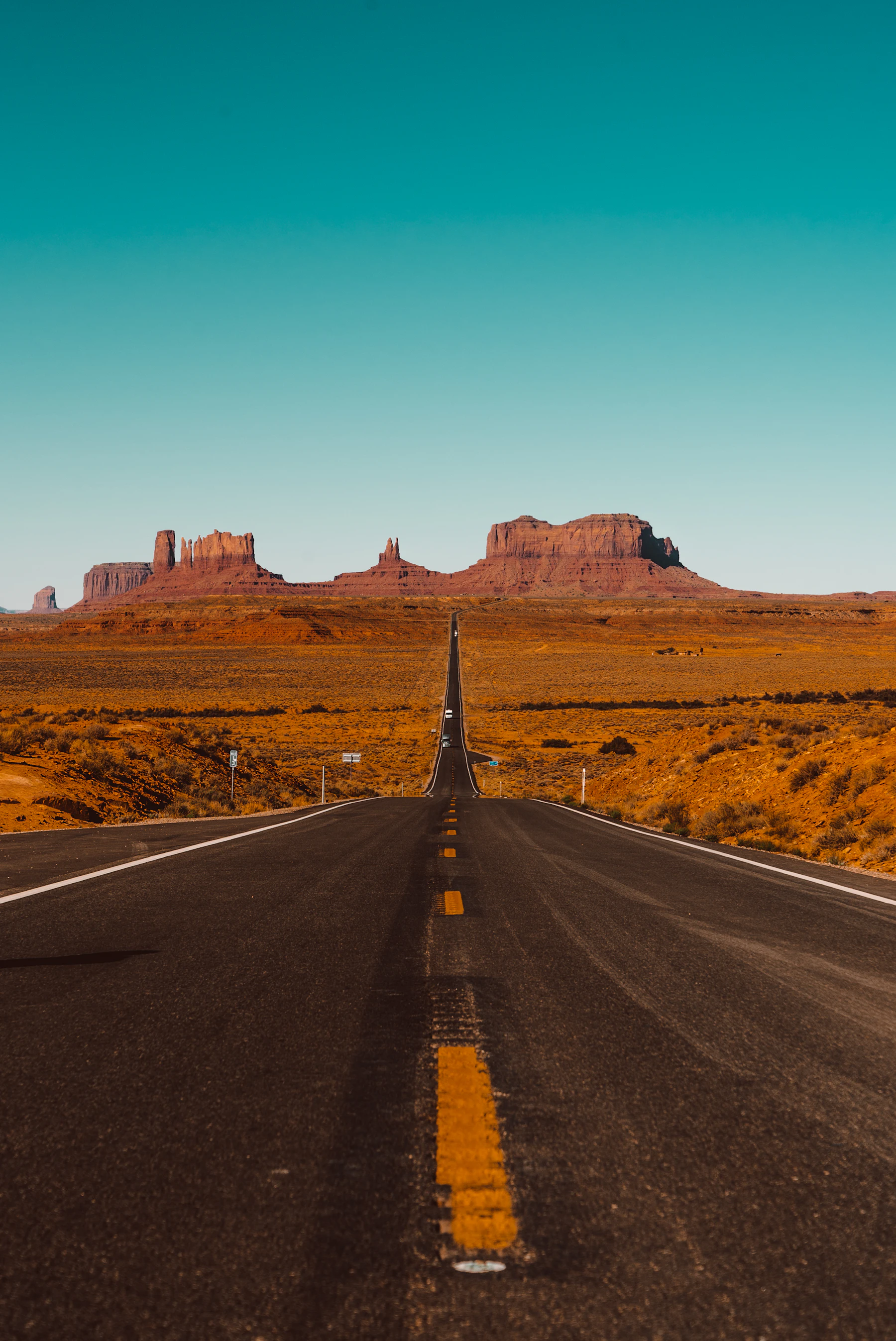 A two-lane American highway running straight toward the Monument Valley sandstone buttes at dusk &mdash; rust earth, asphalt, telephone poles in the distance.
