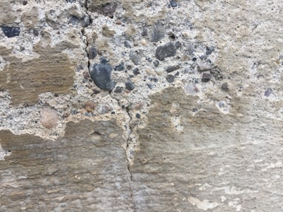 A close-up view of a textured concrete wall with visible cracks and a mix of small stones embedded within it. The surface shows weathering with different shades of gray and beige.
