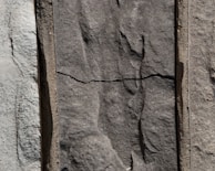 Three sections of vertically arranged stone with varied textures and shades. The center stone has a distinct horizontal crack, giving it an aged appearance. The surface of each stone features natural, rough patterns.