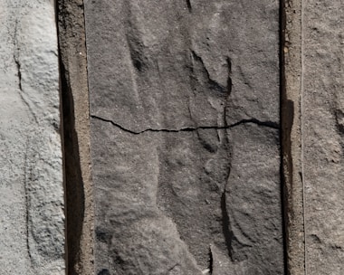 Three sections of vertically arranged stone with varied textures and shades. The center stone has a distinct horizontal crack, giving it an aged appearance. The surface of each stone features natural, rough patterns.