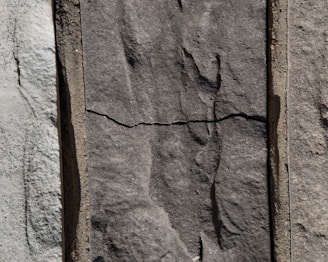 Three sections of vertically arranged stone with varied textures and shades. The center stone has a distinct horizontal crack, giving it an aged appearance. The surface of each stone features natural, rough patterns.