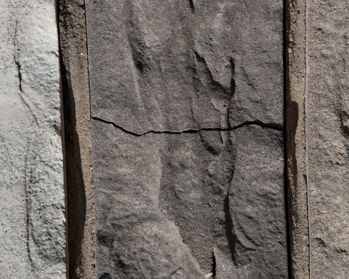 Three sections of vertically arranged stone with varied textures and shades. The center stone has a distinct horizontal crack, giving it an aged appearance. The surface of each stone features natural, rough patterns.