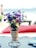 A bouquet of colorful artificial flowers in a woven basket-like holder is placed on a table. The background features a sandy beach and the ocean. A blurred person in swimwear and beach chairs can be seen, indicating a relaxed beach setting.
