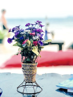 A bouquet of colorful artificial flowers in a woven basket-like holder is placed on a table. The background features a sandy beach and the ocean. A blurred person in swimwear and beach chairs can be seen, indicating a relaxed beach setting.