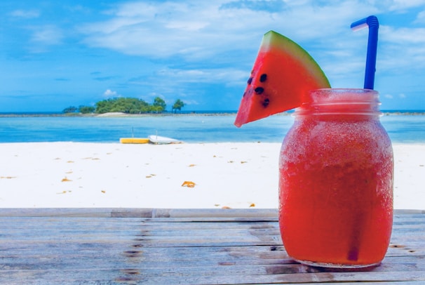 A refreshing watermelon drink in a glass jar with a blue straw sits on a wooden table. A slice of watermelon is wedged on the rim of the jar. In the background, a sandy beach leads to a calm blue ocean, with a small island and two trees visible on the horizon under a partly cloudy sky.