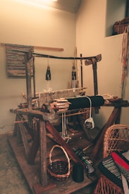 A traditional wooden loom is set up in a small, rustic room. The loom is equipped with various yarns and threads, some of which are woven into a textured fabric on the loom. Surrounding the loom are baskets, chairs, and textiles, suggesting a workspace for weaving. The atmosphere is cozy and homely, with warm lighting creating a vintage feel.