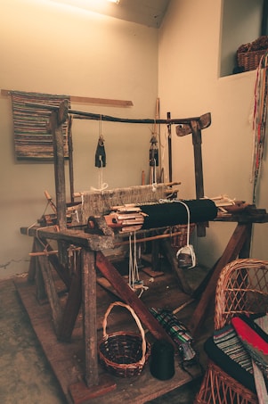 A traditional wooden loom is set up in a small, rustic room. The loom is equipped with various yarns and threads, some of which are woven into a textured fabric on the loom. Surrounding the loom are baskets, chairs, and textiles, suggesting a workspace for weaving. The atmosphere is cozy and homely, with warm lighting creating a vintage feel.