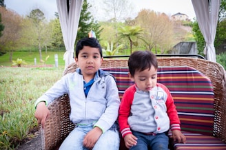 Two young children sit together on a wicker outdoor swing with multicolored cushions in a lush garden setting. The boy on the left wears a light blue jacket and jeans, looking directly at the camera, while the younger child on the right, in a gray and red sweatshirt, looks down. The background features trees and greenery with a glimpse of houses on a hill.