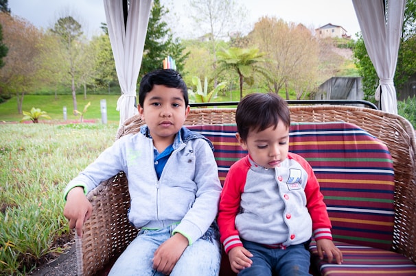 Two young children sit together on a wicker outdoor swing with multicolored cushions in a lush garden setting. The boy on the left wears a light blue jacket and jeans, looking directly at the camera, while the younger child on the right, in a gray and red sweatshirt, looks down. The background features trees and greenery with a glimpse of houses on a hill.