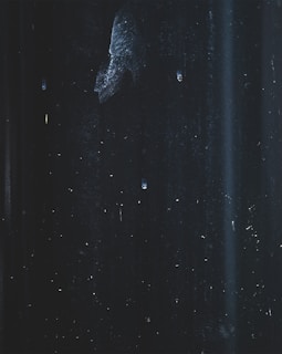 An abstract and dark scene featuring faint streaks and specks, possibly resembling a textured surface or night sky. Subtle variations in light create an impression of depth and mystery.