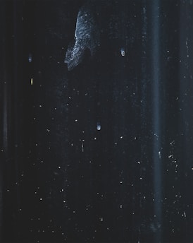 An abstract and dark scene featuring faint streaks and specks, possibly resembling a textured surface or night sky. Subtle variations in light create an impression of depth and mystery.
