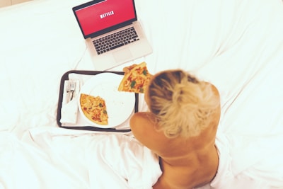 A person sitting on a bed is eating pizza while watching something on a laptop. The laptop screen displays a red background with text. The setting appears relaxed and casual, with white bedding and a tray holding the pizza and some cutlery.
