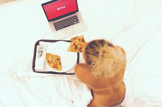 A person sitting on a bed is eating pizza while watching something on a laptop. The laptop screen displays a red background with text. The setting appears relaxed and casual, with white bedding and a tray holding the pizza and some cutlery.