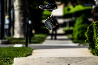 A skateboard mid-air with feet visible above it, performing a trick on a concrete pathway. The background is a blurred green landscape with trees and grass, giving the impression of speed and motion.