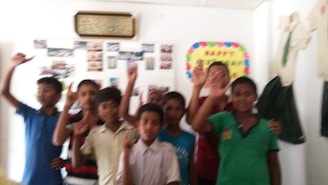 A group of children stands indoors, with some raising their hands and smiling. The wall behind them is decorated with colorful posters and what appear to be photographs. A framed Arabic script is displayed at the top, and school uniform elements can be seen on the right.