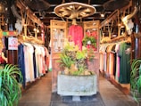 An interior of a clothing store featuring traditional and contemporary garments hanging neatly on racks. The central focus is a decorative wooden structure with intricate carvings, adorned with plants and floral arrangements. The ambiance is warm, with wooden interiors, skulls of cattle mounted on posts, and soft, inviting lighting.