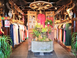 An interior of a clothing store featuring traditional and contemporary garments hanging neatly on racks. The central focus is a decorative wooden structure with intricate carvings, adorned with plants and floral arrangements. The ambiance is warm, with wooden interiors, skulls of cattle mounted on posts, and soft, inviting lighting.