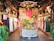 An interior of a clothing store featuring traditional and contemporary garments hanging neatly on racks. The central focus is a decorative wooden structure with intricate carvings, adorned with plants and floral arrangements. The ambiance is warm, with wooden interiors, skulls of cattle mounted on posts, and soft, inviting lighting.