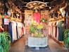 An interior of a clothing store featuring traditional and contemporary garments hanging neatly on racks. The central focus is a decorative wooden structure with intricate carvings, adorned with plants and floral arrangements. The ambiance is warm, with wooden interiors, skulls of cattle mounted on posts, and soft, inviting lighting.