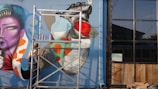 A person on a ladder is creating a large and colorful mural on a wall. The mural depicts a stylized face with purple skin, vibrant orange and white patterns, and a prominent black and red design resembling a feather or hair. The mural's background is primarily blue. Beside the mural, there's a large glass window with reflections and a wooden section beneath it.