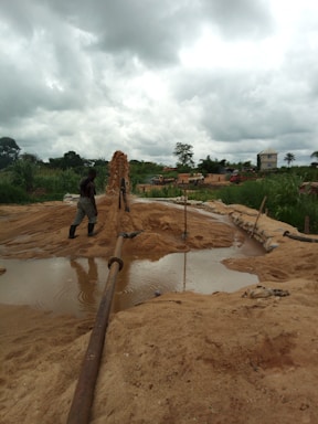 A rural environment with construction activity, featuring a person in boots standing near a large water pipe spewing a stream of sand and water into a basin. The background includes lush greenery, cloudy skies, and several vehicles.