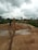 A rural environment with construction activity, featuring a person in boots standing near a large water pipe spewing a stream of sand and water into a basin. The background includes lush greenery, cloudy skies, and several vehicles.