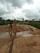 A rural environment with construction activity, featuring a person in boots standing near a large water pipe spewing a stream of sand and water into a basin. The background includes lush greenery, cloudy skies, and several vehicles.