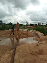 A rural environment with construction activity, featuring a person in boots standing near a large water pipe spewing a stream of sand and water into a basin. The background includes lush greenery, cloudy skies, and several vehicles.