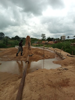 A rural environment with construction activity, featuring a person in boots standing near a large water pipe spewing a stream of sand and water into a basin. The background includes lush greenery, cloudy skies, and several vehicles.