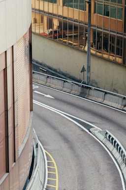 A curved road with lane markings, bordered by concrete barriers and a building. The building has reflective glass windows showing the mirrored image of a red vehicle and urban surroundings. The road is empty and clean, highlighting the cityscape.