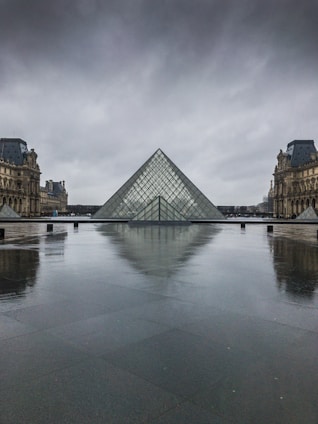 A large glass pyramid set against a dramatic, overcast sky, surrounded by historical architecture with detailed stone carvings. The ground appears wet, reflecting the image of the pyramid and the surrounding buildings. The scene conveys a sense of tranquility and grandeur.