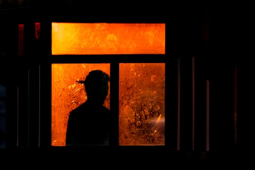 A silhouette of a person appears in front of a window with orange-tinted glass. The glass is weathered and scratched, adding a textured appearance. The scene is dark, with surrounding shadows emphasizing the vibrant orange light coming through the window panes.