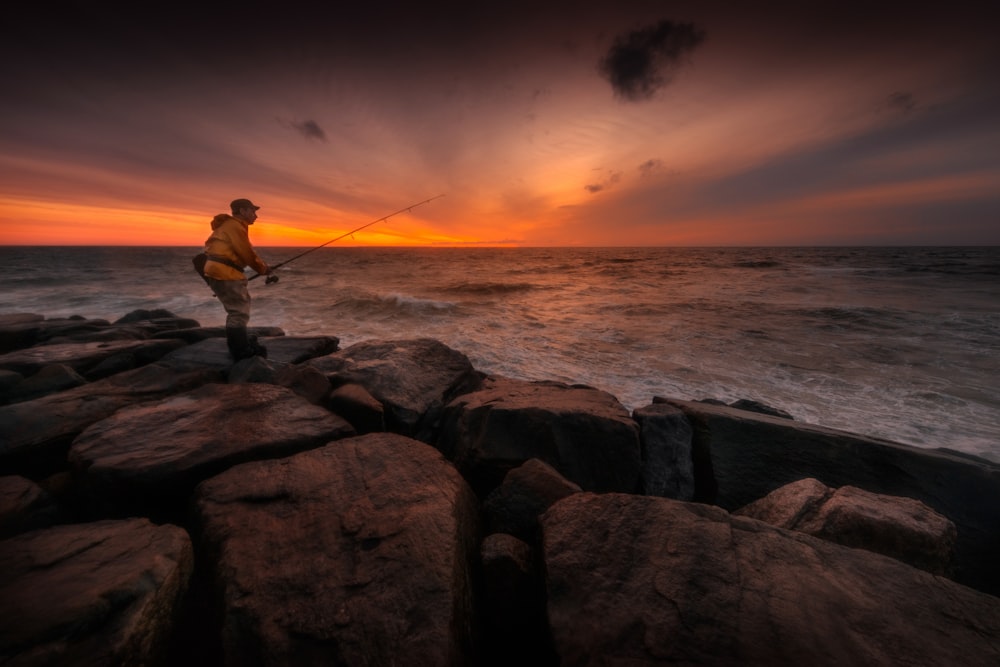 uomo in piedi sulla roccia che pesca sullo specchio d'acqua durante il tramonto