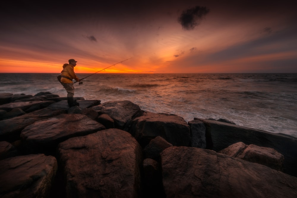 homem em pé na rocha pescando no corpo de água durante o pôr do sol