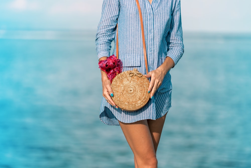 femme portant une robe chemise à rayures bleues et blanches avec un sac à bandoulière en osier marron près de la mer pendant la journée